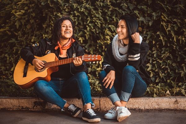 2 junge Frauen sitzen auf Randstein lachen Mädchen spielt Gitarre