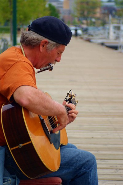 Strassenmusiker spielt sitzend auf Bootssteg Mundharmonika und Gitarre